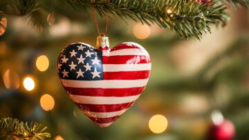 A heart-shaped ornament decorated with the American flag hangs from a Christmas tree, surrounded by festive holiday lights and bokeh.