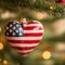 A heart-shaped ornament decorated with the American flag hangs from a Christmas tree, surrounded by festive holiday lights and bokeh.