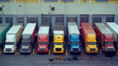 Fleet of colorful semi trucks are parked in front of loading docks at a large distribution warehouse building waiting to be unloaded