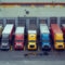 Fleet of colorful semi trucks are parked in front of loading docks at a large distribution warehouse building waiting to be unloaded